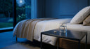 A minimalist nighttime bedroom scene with cool blue ambient lighting, crisp white bedding, and a clean nightstand with a glass of water and a single wellness capsule.
